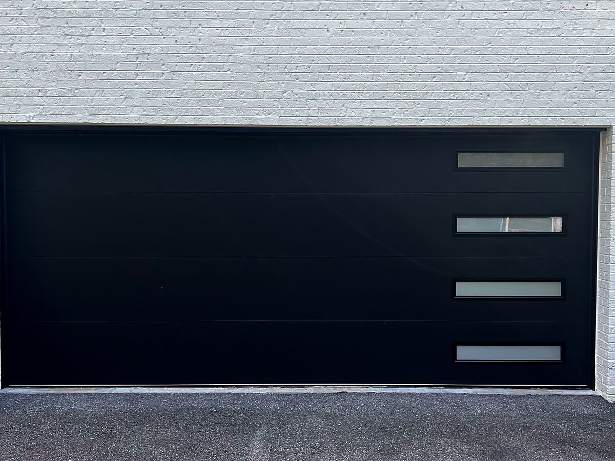 Modern black flush garage door with a column of windows on a white brick home in Garrison